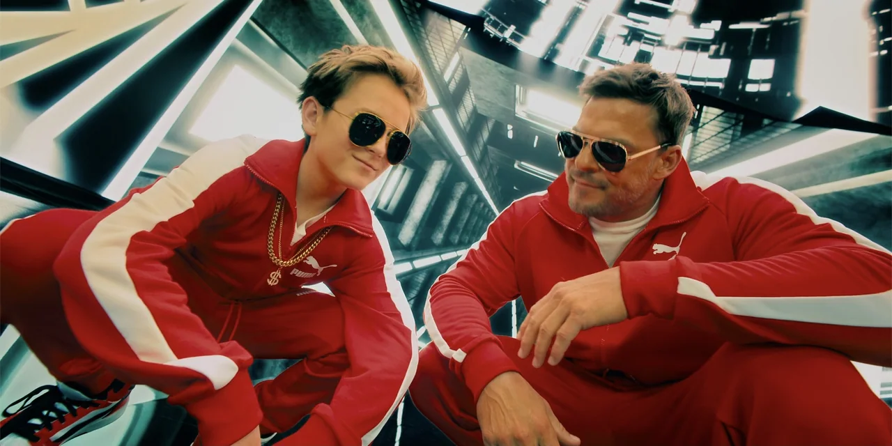 Playdate (2025) movie still: medium — A modern corridor features sharp geometric lines and bright white paneling. Two men in matching red athletic…
