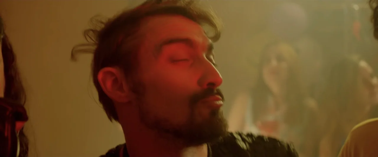 Perdida (2018) movie still: closeup — A crowded interior party space with haze and blurred figures in the background. A man with a dark beard dances…