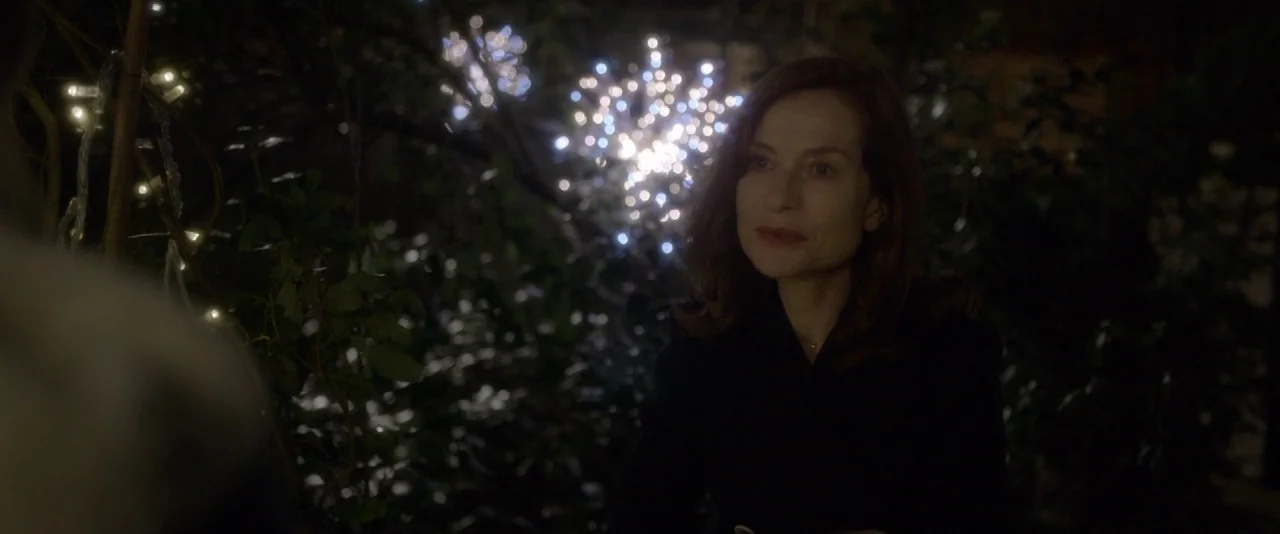 Elle (2016) movie still: medium — A woman stands outdoors at night in front of dark, dense foliage. Clusters of bright white LED string lights hang…