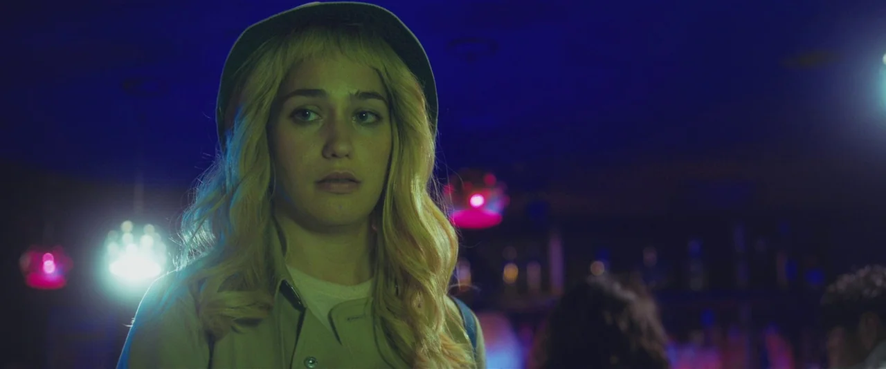 Gemini (2017) movie still: medium — A dimly lit bar interior filled with vibrant neon colored light sources. A young woman with blonde hair stands…