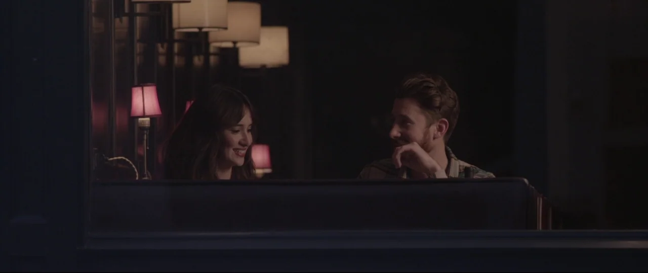 Sun Choke (2015) movie still: medium — Two people sit together in a booth inside a dark, quiet establishment at night. A young woman and a bearded…