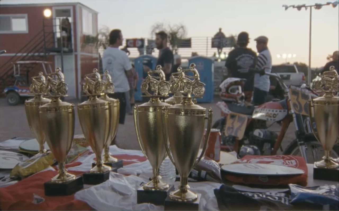 The Greasy Hands Preachers (2014) movie still: medium — A row of gold motorcycling trophies sits on a table outdoors at a dirt track. Several people…