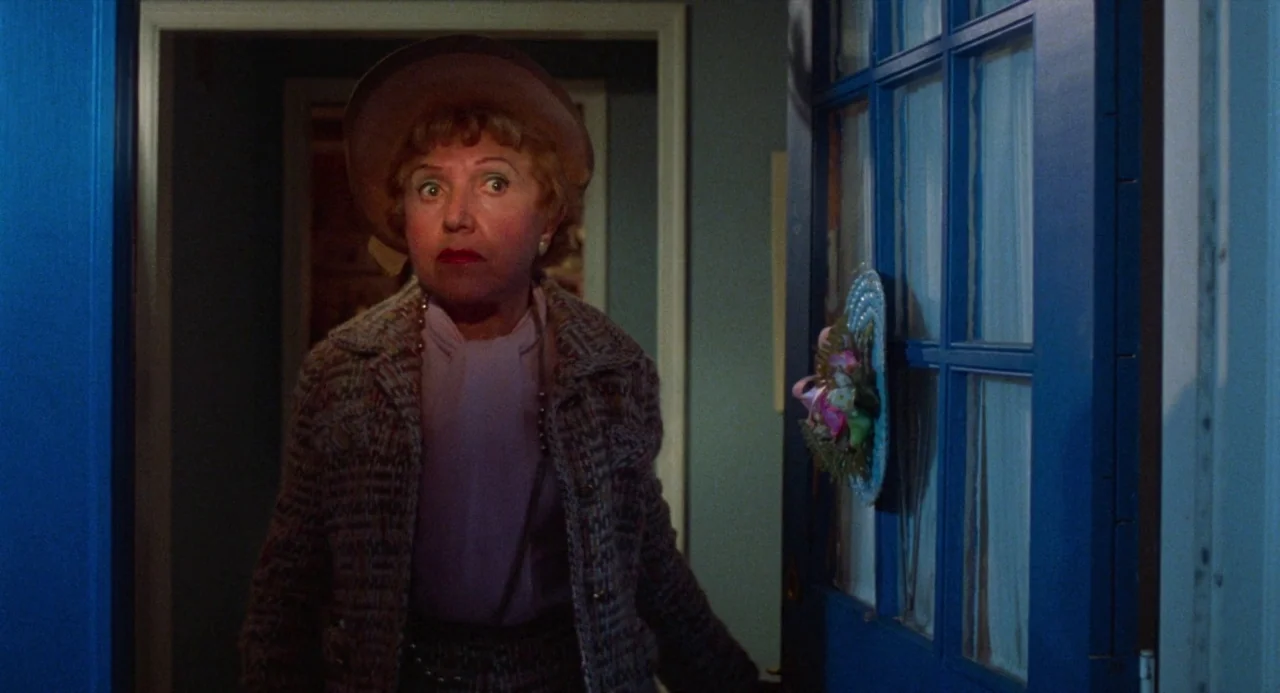 Polyester (1981) movie still: medium — An interior hallway features a blue door frame and a plain grey wall background. A woman in a tweed jacket…