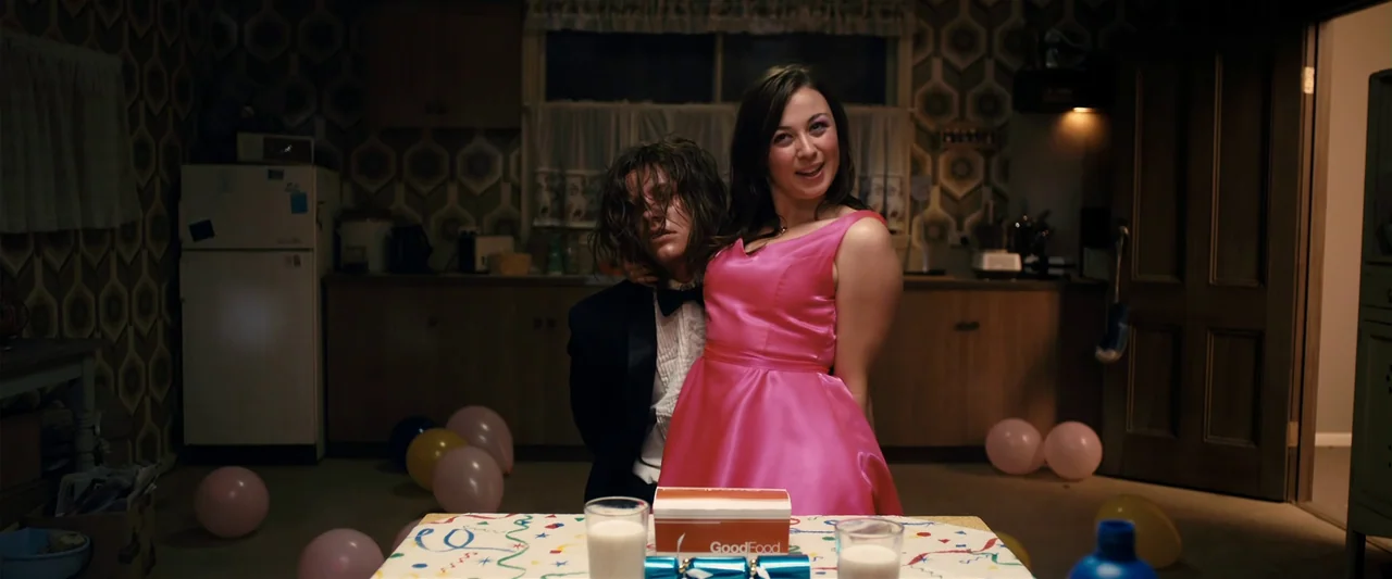The Loved Ones (2009) movie still: medium — A dark dining room features patterned wallpaper and balloons scattered on the floor. A woman in a pink…