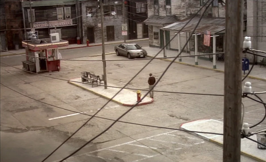 Dear Wendy (2005) movie still: wide — A quiet, paved parking lot features a small food kiosk and surrounding aged buildings. A man stands alone in…