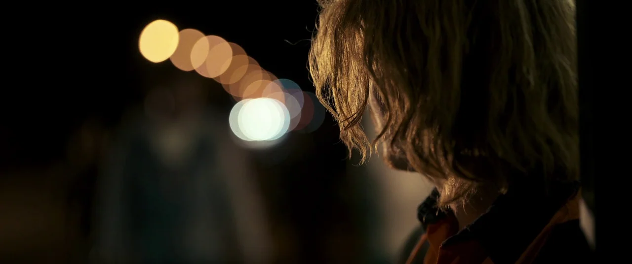 Talk To Me (2022) movie still: closeup — A person stands at night next to a dark area with bokeh lights. The subject is backlit by a warm source…