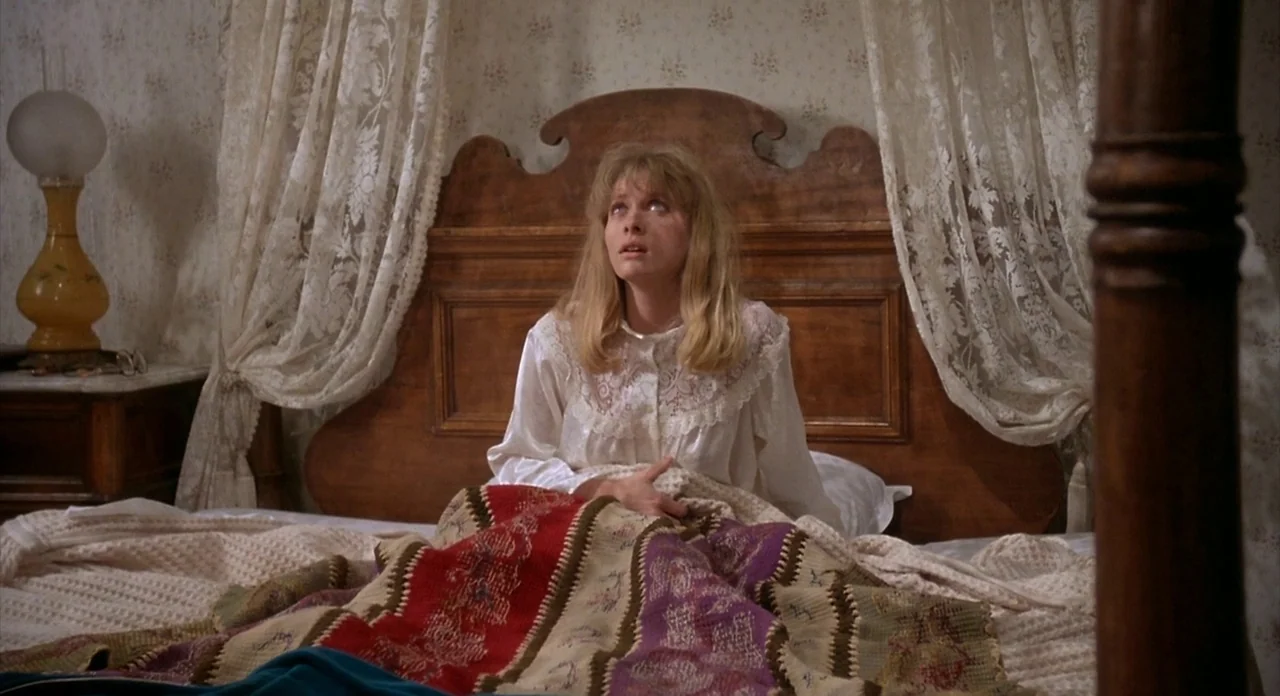 From Beyond (1986) movie still: medium — A woman sits upright in a bed with a wooden headboard and lace curtains. She grips a colorful knit blanket…