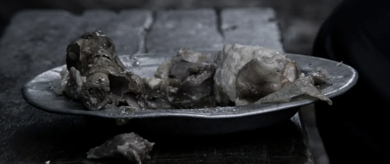 Van Diemens Land (2009) movie still: closeup — A metal plate holding pieces of cooked meat sits on a weathered wooden table. The plate appears worn…