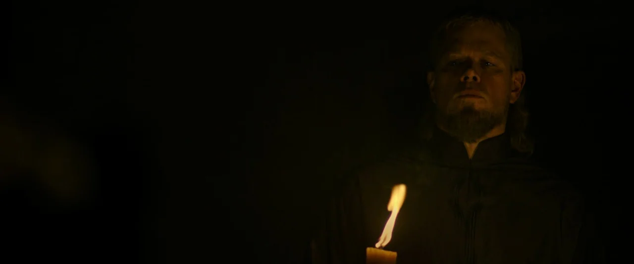 The Last Duel (2021) movie still: medium — A dark, undifferentiated interior space is shrouded in deep shadow. A man holds a single lit candle that…