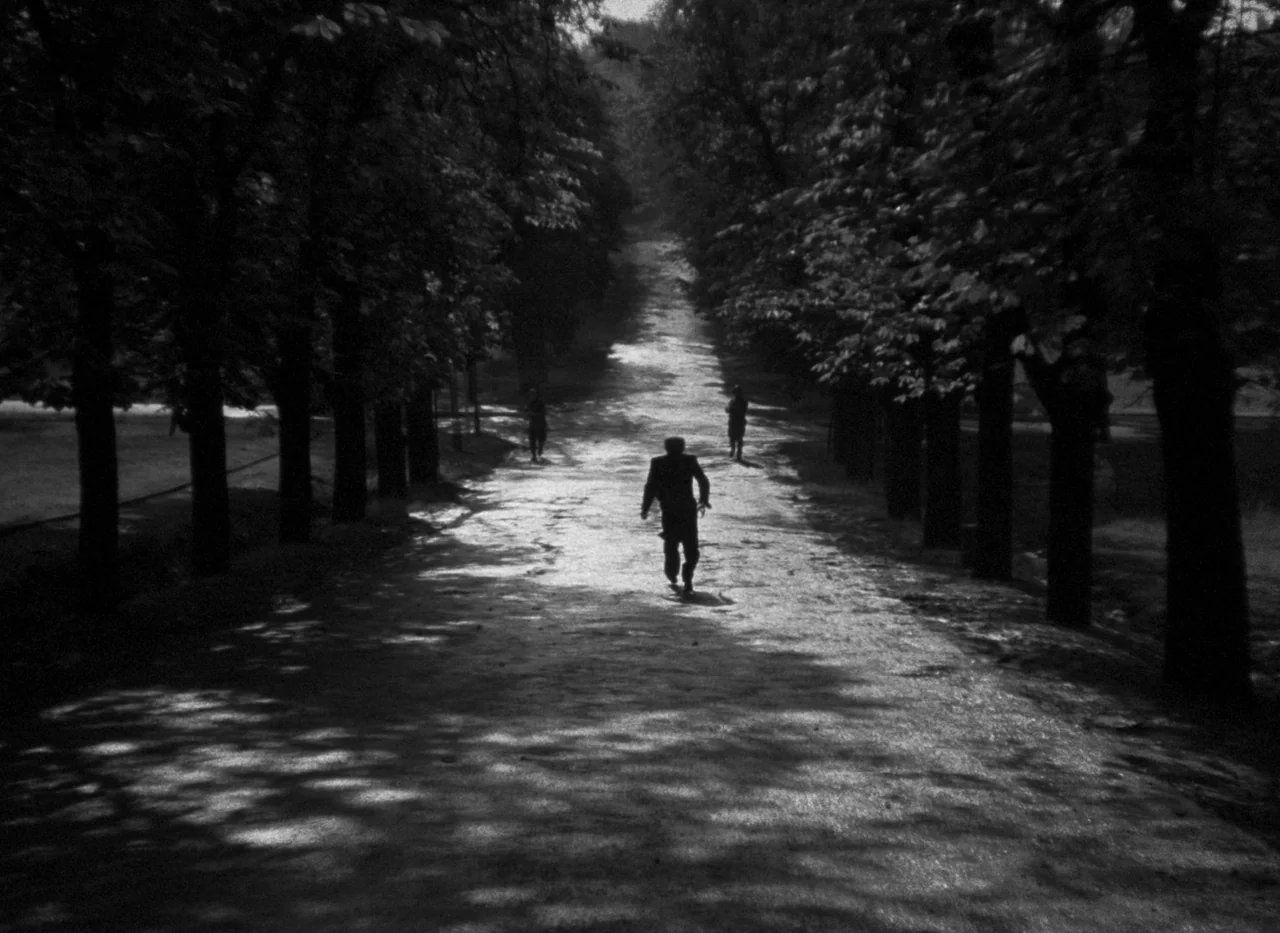 Krakatit (1948) movie still: wide — A long, dirt path stretches into the distance, framed by rows of tall, dark trees. A single figure walks down…