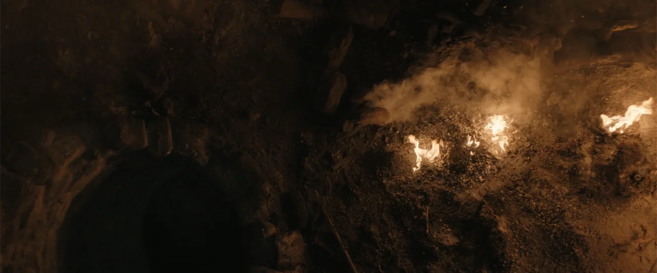The Bluff (2026) movie still: establishing — An uneven dirt surface features small flames burning amidst scattered rocks and debris. A dark, hollow…