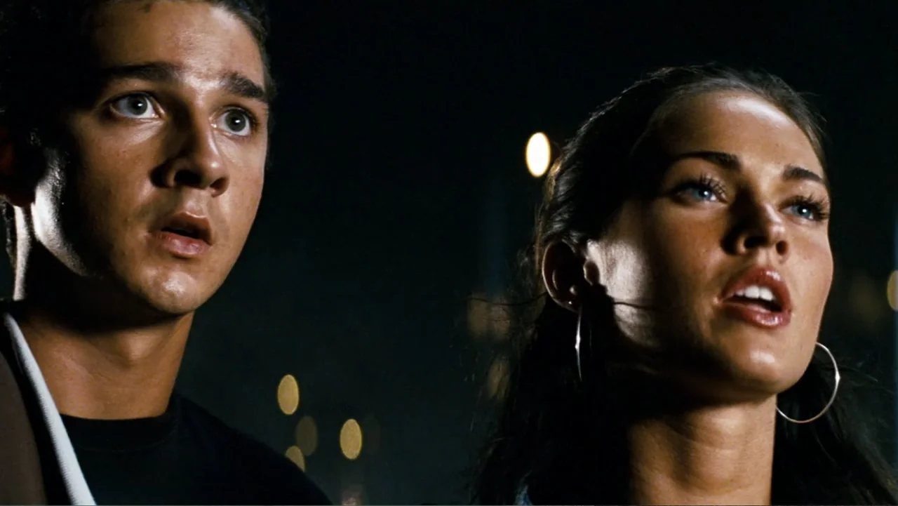 Transformers (2007) movie still: closeup — Two individuals stand against a dark night background with out-of-focus city lights behind. The man and…
