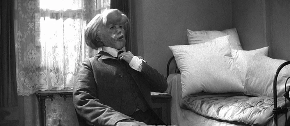 The Elephant Man (1980) movie still: medium — An interior space features a bed piled with multiple white pillows against a wall. A man with…