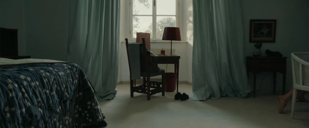 Peacock (2010) movie still: wide — A furnished bedroom features a writing desk placed directly in front of a window. Light curtains hang on both…