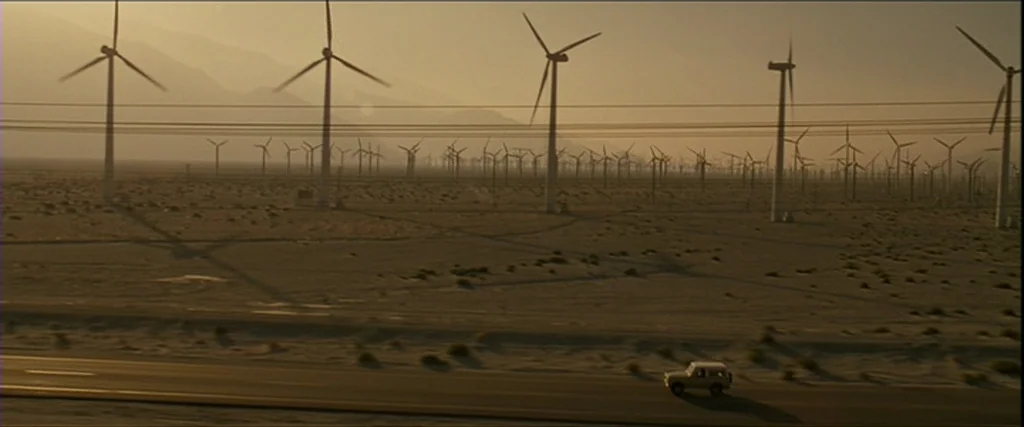 Into the Wild (2007) movie still: wide — A vast, flat desert landscape is filled with numerous tall white wind turbines. A small vehicle travels…