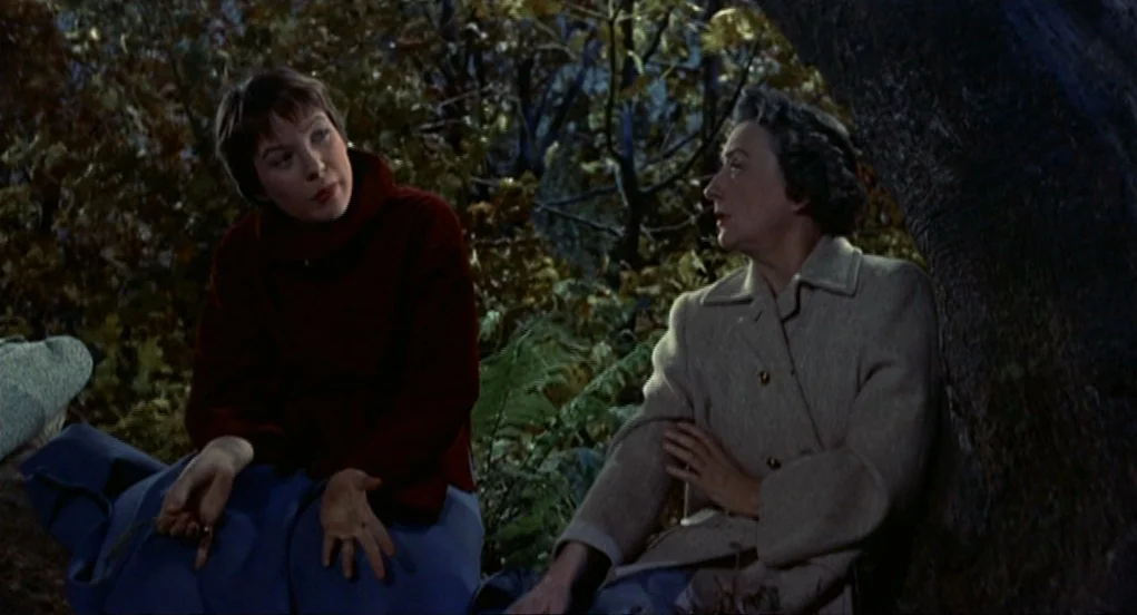 The Trouble With Harry (1955) movie still: medium — Two women sit together in a dense wood during the night time hours. They are positioned near the…