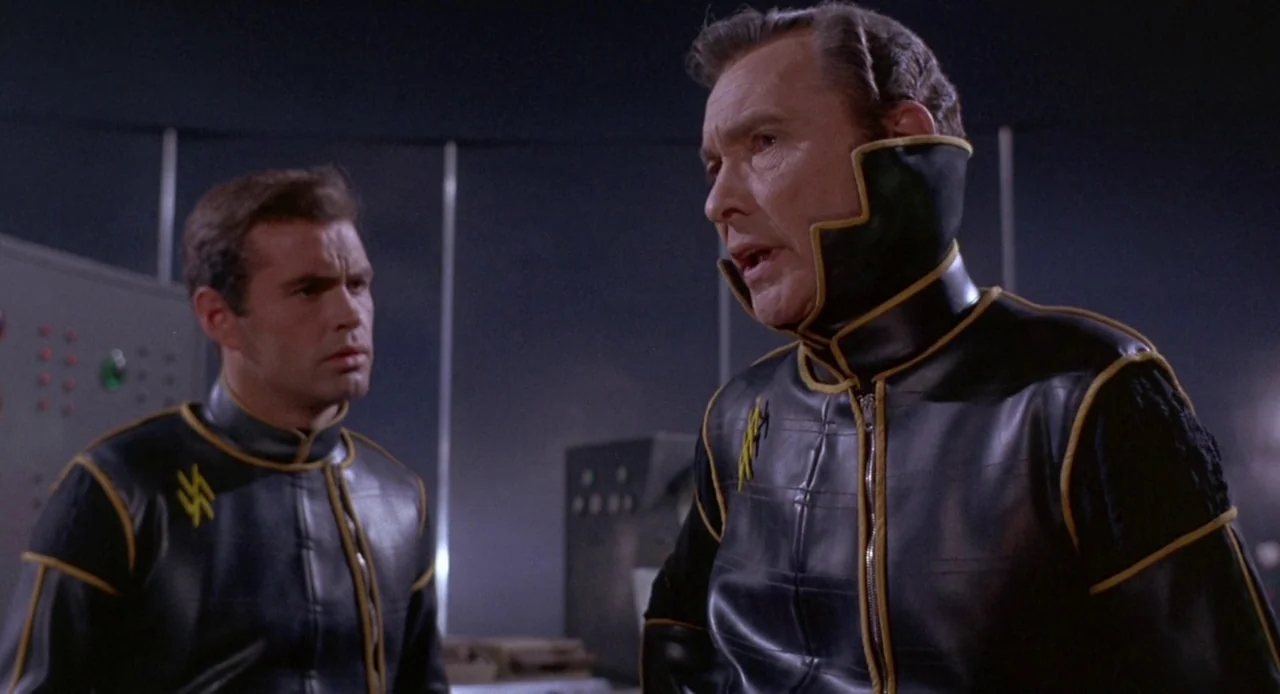 Planet of the Vampires (1965) movie still: medium — A dark technical room contains metallic control panels and grey equipment in the background. Two…