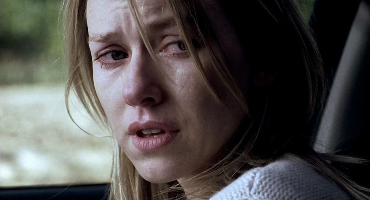 21 Grams (2003) movie still: closeup — A woman sits inside a car with a view of greenery through the window. She looks toward the camera with…