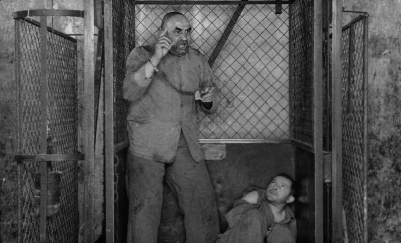 Goto, Island of Love (1969) movie still: fullbody — A man stands inside a metal wire cage enclosure while another man sits below. The enclosure is…