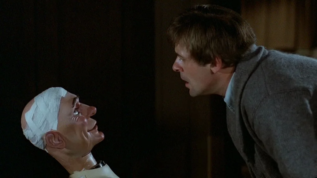 Magic (1978) movie still: medium — A dimly lit room provides the setting for a interaction between two figures. A man leans closely towards a…