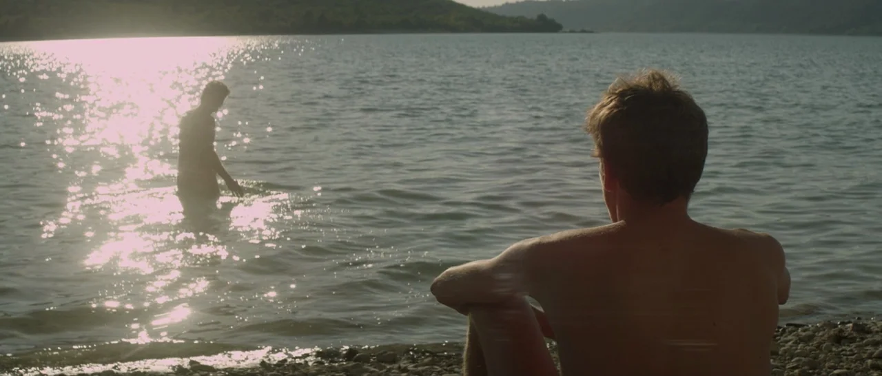 Stranger By The Lake (2013) movie still: medium — A rocky lakeshore stretches toward distant low hills under a bright clear sky. One man sits in the…
