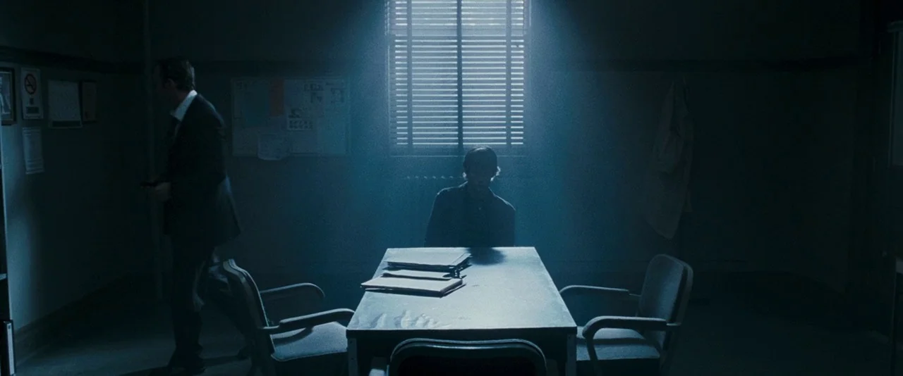 Dead Silence (2007) movie still: wide — A dimly lit room features a metal table centered beneath a window with blinds. Two men are inside the space…