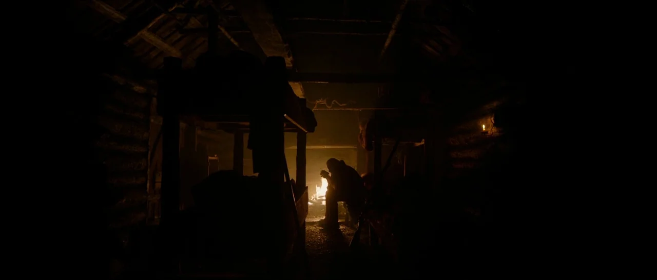 The Revenant (2015) movie still: wide — A dark log cabin interior features wooden walls and heavy timber framing beams. A solitary man sits near a…