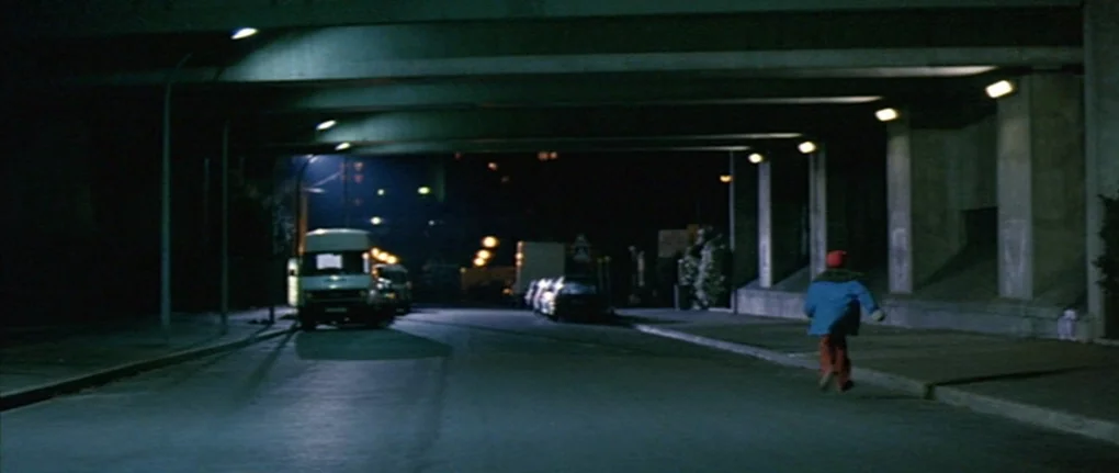 IP5 (1992) movie still: wide — A dark concrete underpass at night featuring repeating pillars and overhead road structure. A small child in a blue…
