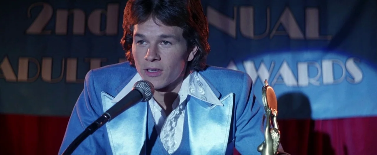 Boogie Nights movie still: medium — A man stands in front of a banner announcing an annual adult awards ceremony. He wears a light blue satin tuxedo…