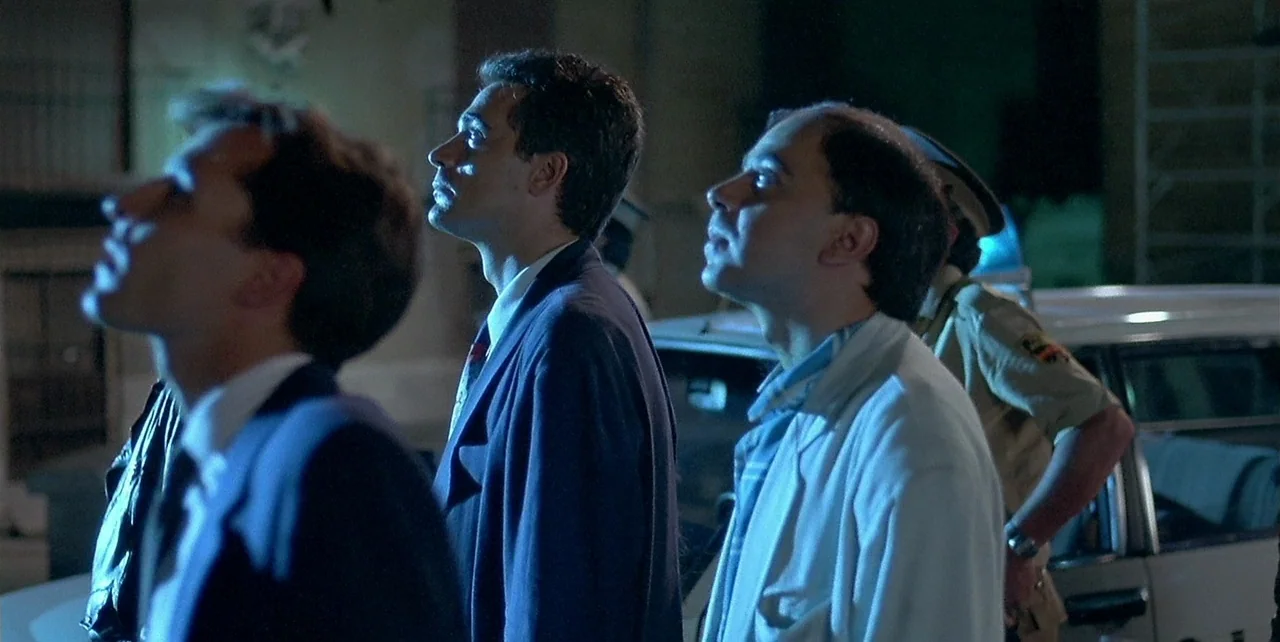 Law of Desire (1987) movie still: medium — A nighttime parking lot exterior features several men standing next to a parked car. The group gazes…