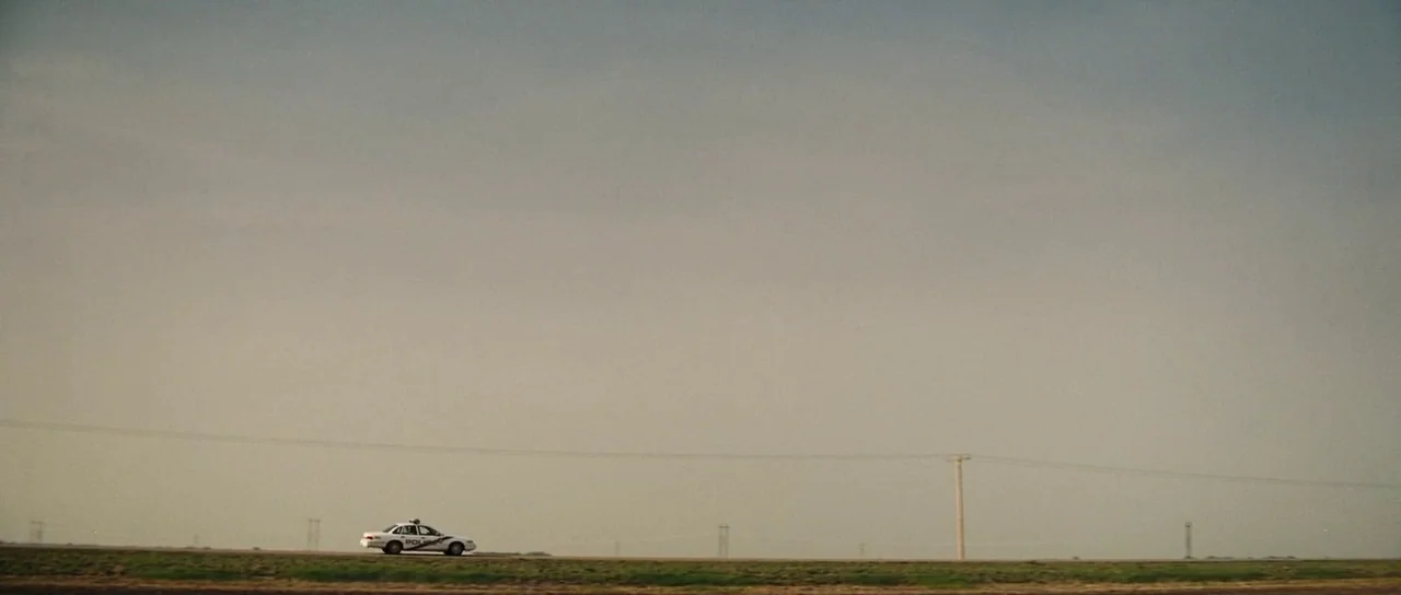 Surveillance (2008) movie still: establishing — A desolate, flat landscape features a single road stretching across the horizon. A white police car…