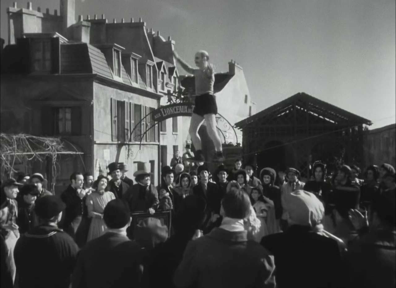 Les Enfants Du Paradis (1945) movie still: wide — An open town square features European-style buildings and an arched sign reading Aux Lionceaux. A…