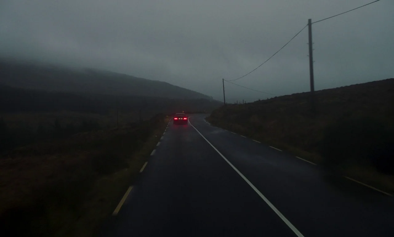 It Is In Us All (2022) movie still: wide — An empty asphalt road stretches through a hilly landscape under thick grey clouds. A single vehicle with…