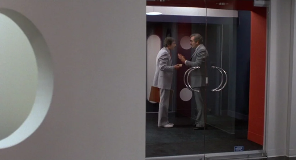 The King Of Comedy (1982) movie still: medium — An office entrance with glass double doors is framed by plain white and red walls. Two men in…