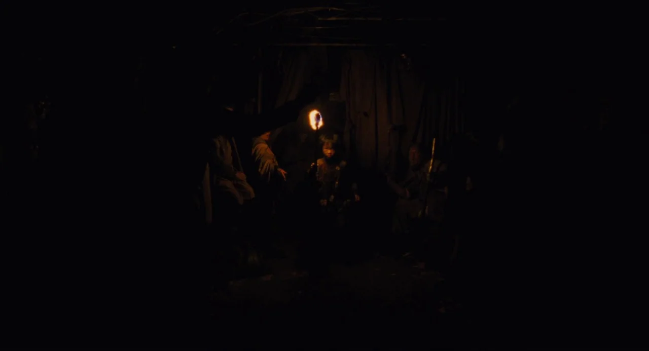 Snowpiercer (2013) movie still: medium — A group of people sits in a dark, makeshift shelter with fabric walls. A child stands in the center holding…