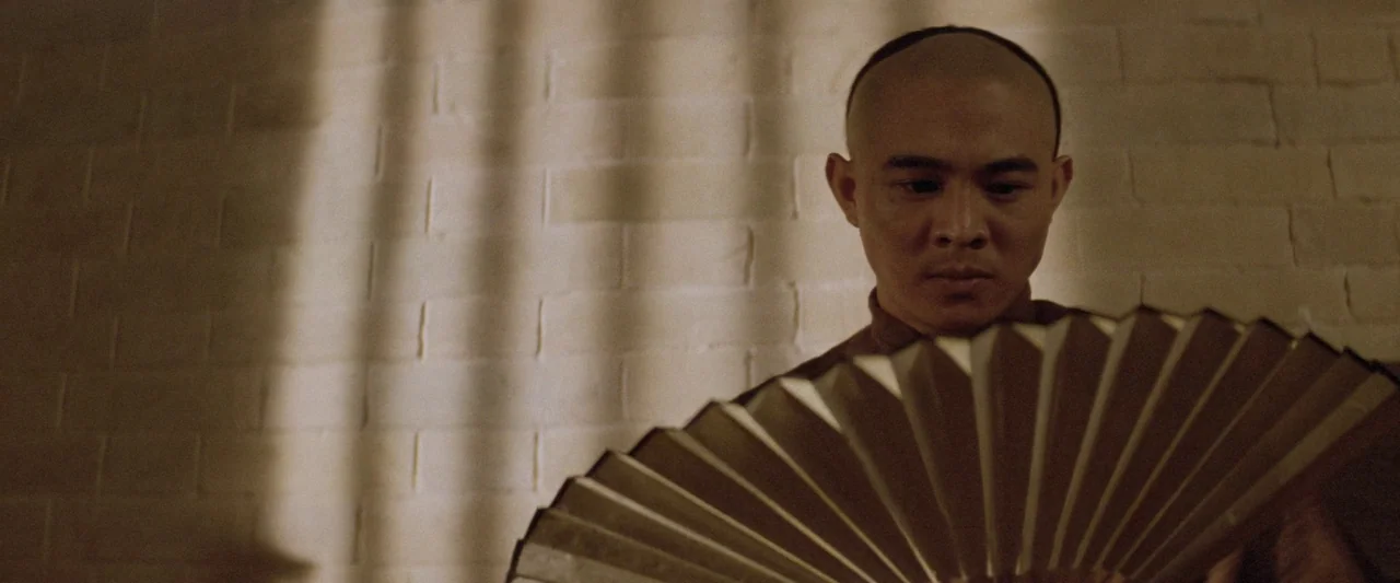 Once Upon a Time in China (1991) movie still: closeup — A man stands before a brick wall textured with long vertical shadow bands. He holds a large,…