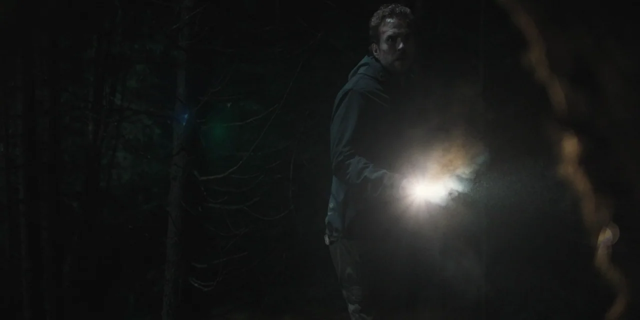 The Ritual (2017) movie still: medium — A dense dark forest at night with tall trees and thick vegetation. A man in a dark jacket holds a flashlight…