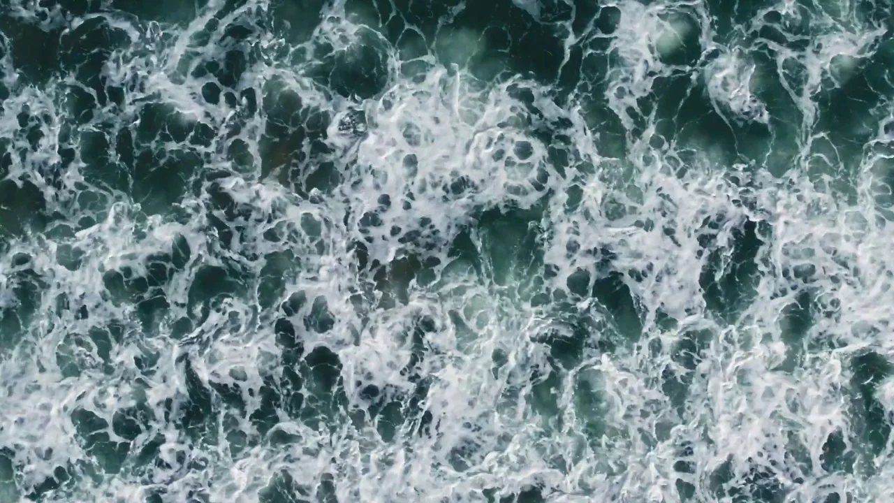 Black Mother (2018) movie still: establishing — A top-down view shows the surface of the ocean with turbulent white sea foam. The waves overlap and…
