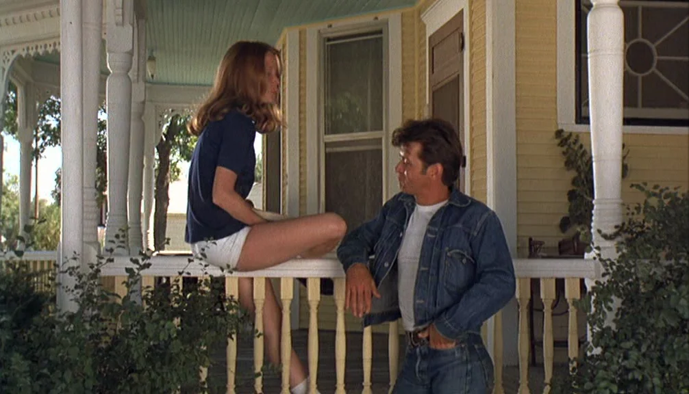 Badlands (1973) movie still: fullbody — A wooden porch of a yellow house with painted spindles and white trim. A young woman sits on the railing as…