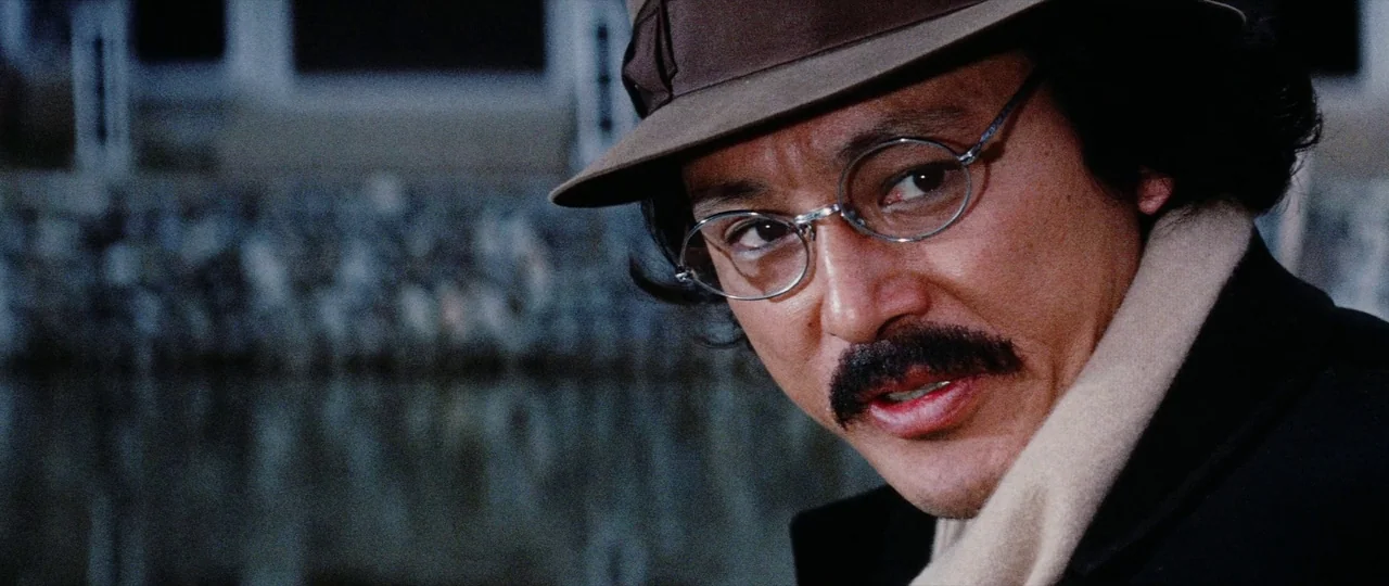 Lady Snowblood Song of Vengeance (1974) movie still: closeup — A man in a fedora stands outdoors near a water-covered structure or dock. He looks…