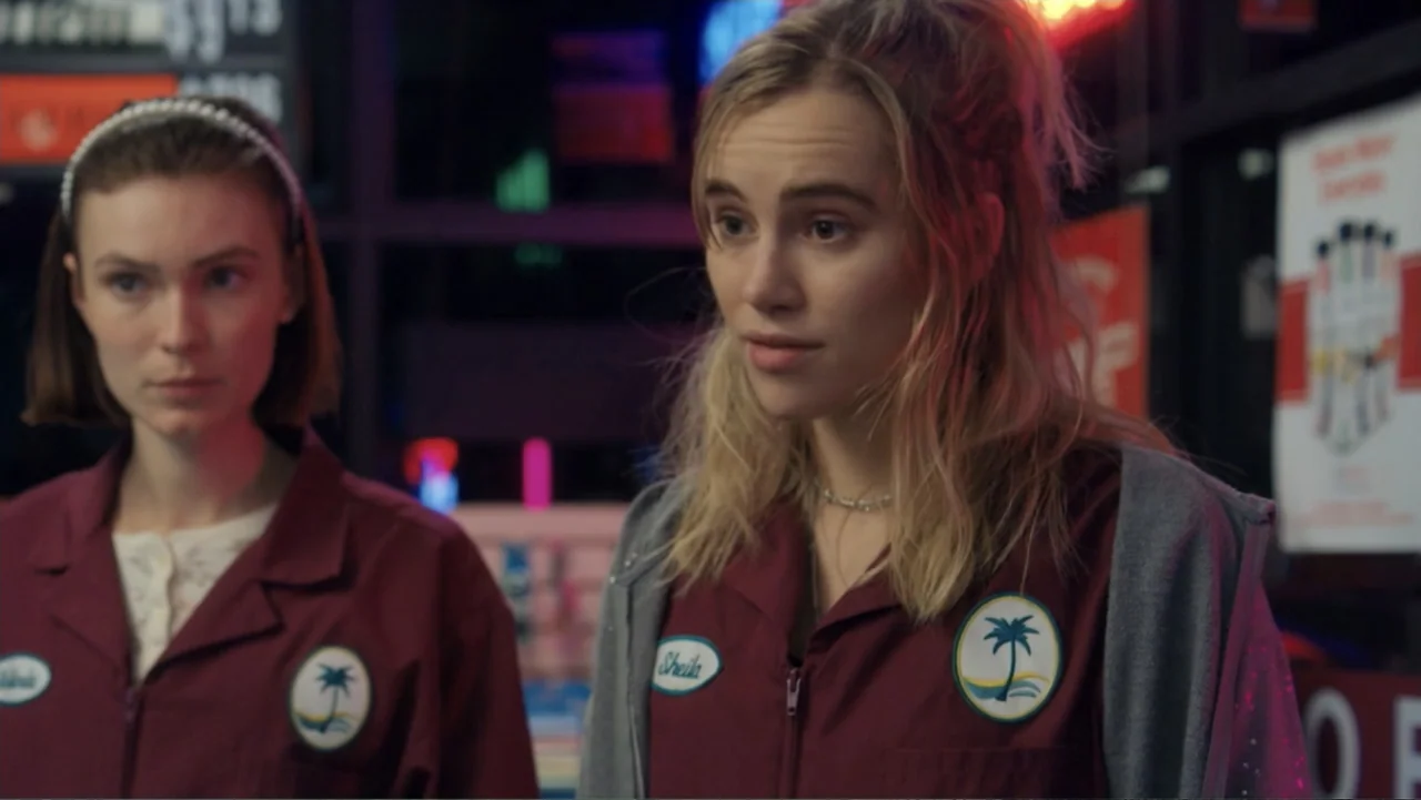 Burn (2019) movie still: medium — Two young women in work uniforms stand indoors against a background of shelves. They are wearing matching maroon…