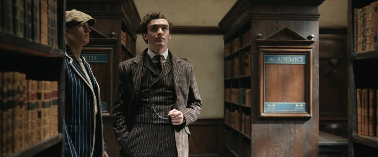 Young Sherlock Holmes (1985) movie still: medium — A historic library features tall wooden shelving units filled with leather-bound volumes. Two men…