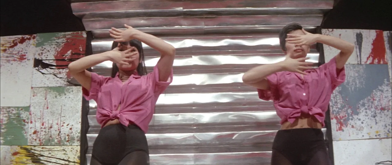 Black Tight Killers (1966) movie still: medium — A performance area features a crinkled metal wall flanked by panels with paint splatter. Two women…