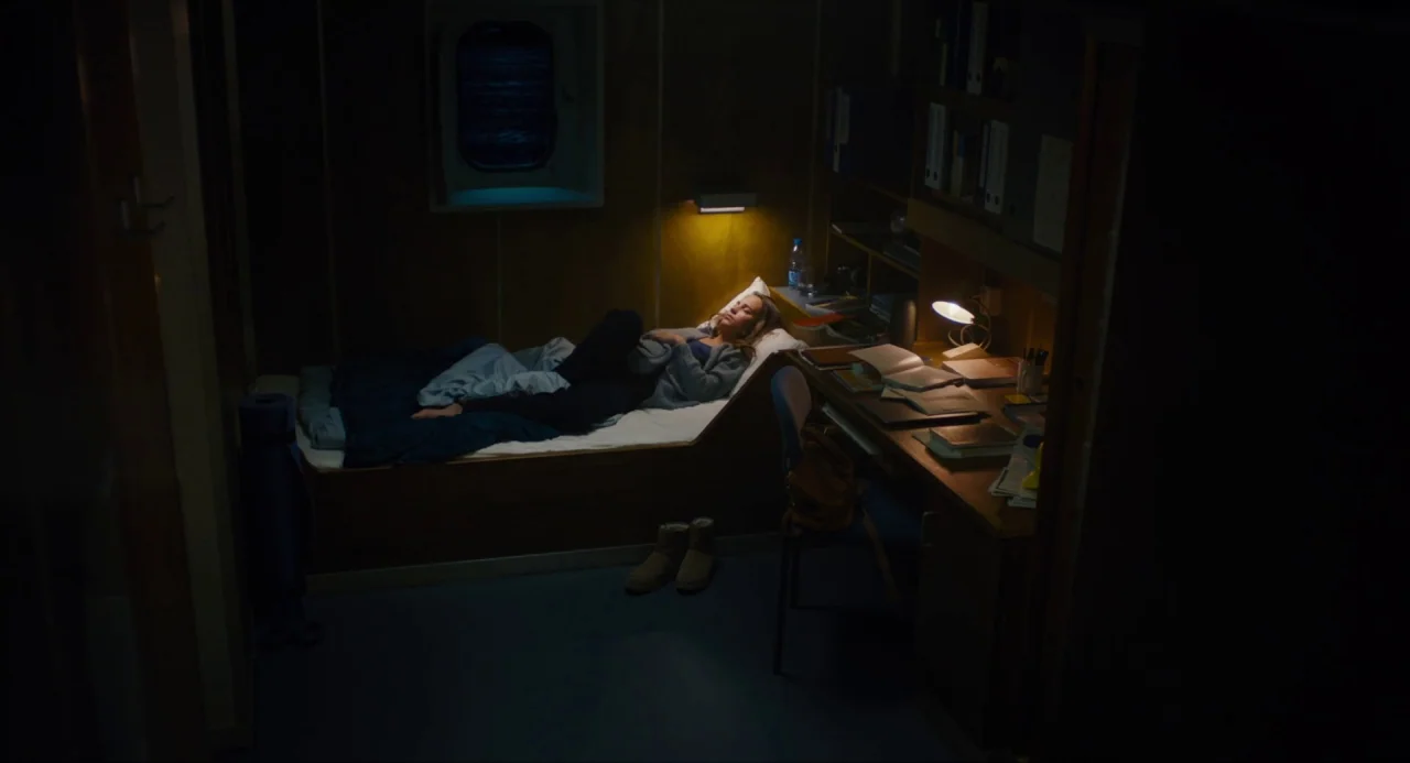 Submergence movie still: wide — A dimly lit, cramped cabin on a vessel features wooden walls and a porthole. A woman rests on a single bed next to a…