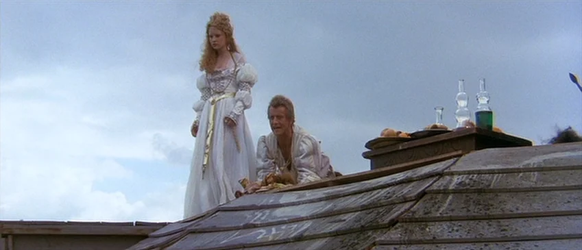 Flesh+Blood (1985) movie still: fullbody — A man and a woman in historical fantasy attire stand on a tiled rooftop. A wooden tray with glass bottles…