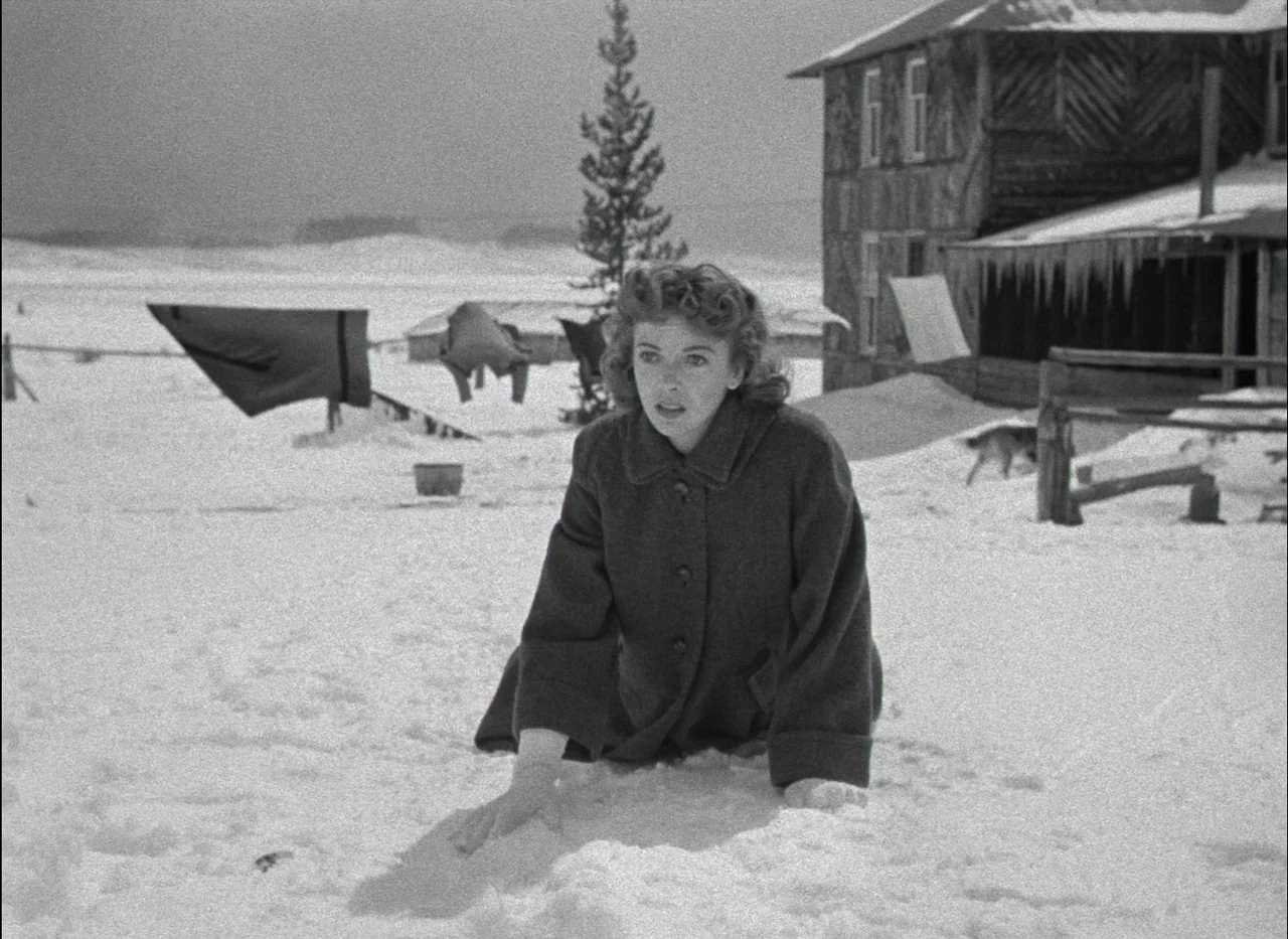 On Dangerous Ground (1951) movie still: medium — A woman crawls across a vast snow-covered field in front of a rustic wooden building. Laundry hangs…