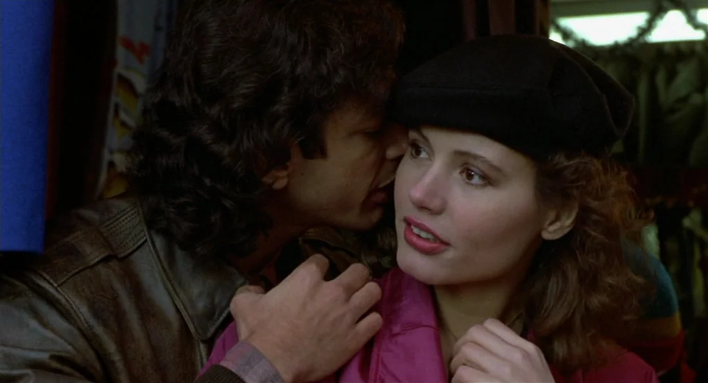 The Fly (1986) movie still: closeup — A dimly lit interior space with dark textures and clothing visible in the background. A man leans in close to…