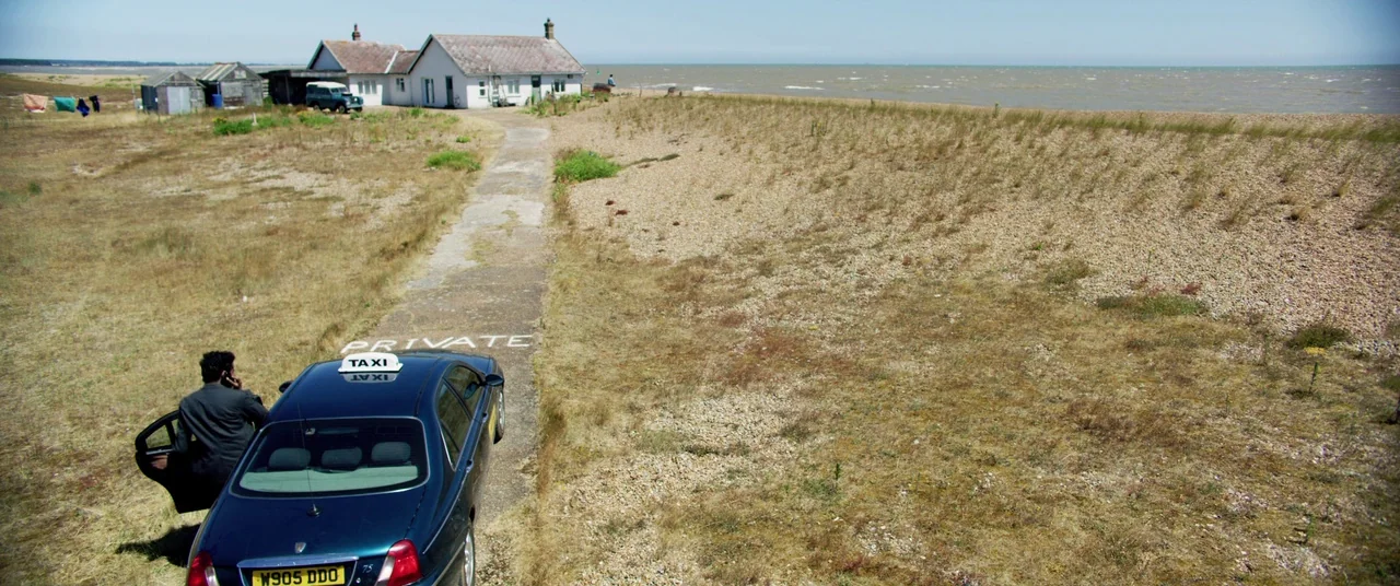 Yesterday (2019) movie still: wide — A gravel-lined coastal path leads toward small houses and the open sea. A man in a dark suit stands beside a…
