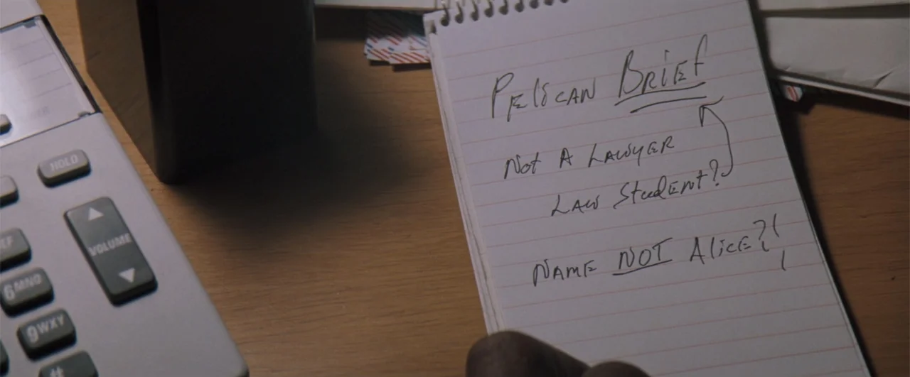 The Pelican Brief (1993) movie still: closeup — A close-up of a white notepad with handwritten text sits on a wooden desk. A portion of a telephone…