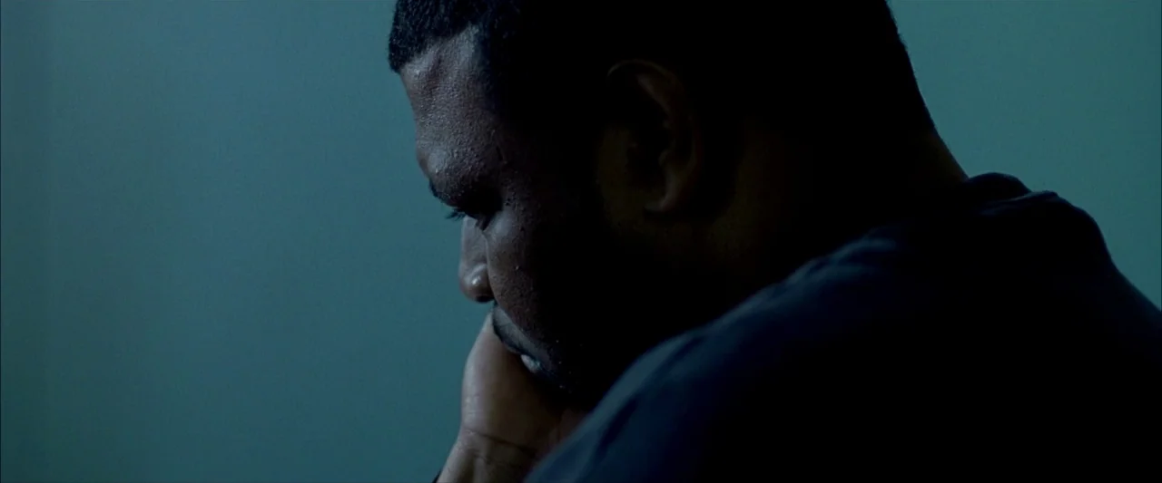 Ballast (2008) movie still: closeup — A dark, sparsely lit indoor space with a solid blue-toned wall as background. A man sits in profile, resting…